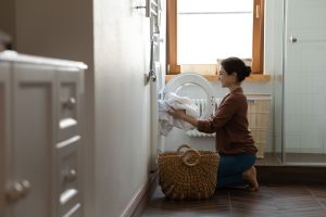 A woman kneeling on the floor is seen loading white laundry into a front-loading washing machine. She has a woven basket beside her, placed on dark wooden flooring. The room is softly lit by natural light from a nearby window, with a simple, tidy setting that includes a laundry hamper and tiled walls. The scene conveys a peaceful, domestic atmosphere.