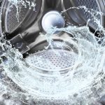 A close-up view of the inside of a washing machine drum with water splashing in dynamic motion. The shiny, perforated metal interior reflects light, while the water appears to swirl and splash, capturing the action of a wash cycle. The image highlights the stainless steel texture and the fluidity of water in the machine.