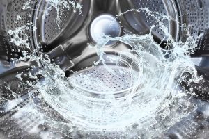 A close-up view of the inside of a washing machine drum with water splashing in dynamic motion. The shiny, perforated metal interior reflects light, while the water appears to swirl and splash, capturing the action of a wash cycle. The image highlights the stainless steel texture and the fluidity of water in the machine.