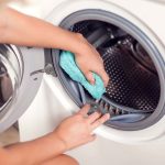 A person is cleaning the rubber seal of a front-loading washing machine using a turquoise cloth. The open door reveals the shiny metal drum, and the person's hands are focused on wiping the inner gasket area to ensure cleanliness. The scene emphasizes maintenance and care for household appliances.