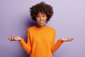 A young woman with curly hair stands against a purple background, wearing an orange sweater. She has a confused or questioning expression, with her eyebrows raised and one side of her mouth slightly twisted. Her arms are extended outward with palms up, as if asking, "What?" or showing uncertainty.