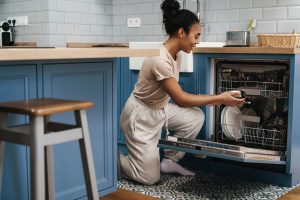 A woman kneels in front of an open dishwasher in a modern kitchen with blue cabinets and patterned tile flooring. She is loading or unloading dishes, including plates and bowls, and is dressed casually in a beige t-shirt and gray sweatpants. The countertop holds a small appliance, a basket, and cleaning supplies.