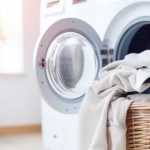 A laundry room scene with a wicker basket filled with folded towels and linens in front of an open clothes dryer machine. Sunlight filters softly through a nearby window, illuminating the room and casting a warm glow over potted plants in the background.