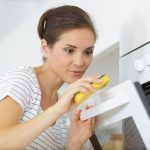 A focused woman cleaning the door of a white oven using a yellow sponge. She is wearing a white shirt with thin black stripes, and her hair is tied back. The scene is bright and minimalistic, with the oven controls visible on the side.