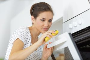 A focused woman cleaning the door of a white oven using a yellow sponge. She is wearing a white shirt with thin black stripes, and her hair is tied back. The scene is bright and minimalistic, with the oven controls visible on the side.