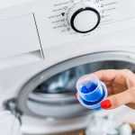 A close-up of a person’s hand with red nail polish holding a small measuring cap filled with blue liquid laundry detergent in front of a front-loading washing machine. The washing machine's control panel is visible, showing various wash cycle options. The open door reveals laundry inside.