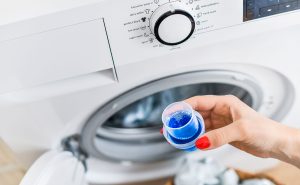 A close-up of a person’s hand with red nail polish holding a small measuring cap filled with blue liquid laundry detergent in front of a front-loading washing machine. The washing machine's control panel is visible, showing various wash cycle options. The open door reveals laundry inside.