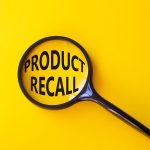 A black magnifying glass placed on a yellow background, enlarging the bold black text "PRODUCT RECALL" within its lens.