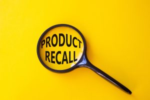 A black magnifying glass placed on a yellow background, enlarging the bold black text "PRODUCT RECALL" within its lens.