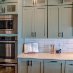 A modern kitchen with sage green cabinets, black handles, and a white subway tile backsplash. A stainless steel microwave and double oven are built into the cabinetry. The quartz countertop features an open cookbook on a wooden stand, wooden salt and pepper grinders, and a mortar and pestle. A gas stovetop with a wooden cutting board is partially visible on the right. The kitchen has a clean and organized appearance with warm, natural lighting.