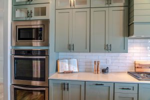 A modern kitchen with sage green cabinets, black handles, and a white subway tile backsplash. A stainless steel microwave and double oven are built into the cabinetry. The quartz countertop features an open cookbook on a wooden stand, wooden salt and pepper grinders, and a mortar and pestle. A gas stovetop with a wooden cutting board is partially visible on the right. The kitchen has a clean and organized appearance with warm, natural lighting.