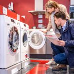 A man and a woman are shopping for washing machines in an appliance store. The man, wearing a navy blue jacket and jeans, is crouching while holding an information sheet. The woman, wearing a beige coat, is leaning over and examining the open door of a front-loading washing machine. The store has red walls and a modern design, with other appliances visible in the background.