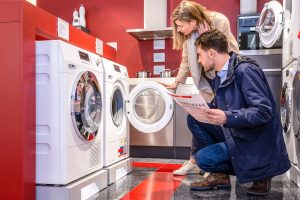 A man and a woman are shopping for washing machines in an appliance store. The man, wearing a navy blue jacket and jeans, is crouching while holding an information sheet. The woman, wearing a beige coat, is leaning over and examining the open door of a front-loading washing machine. The store has red walls and a modern design, with other appliances visible in the background.