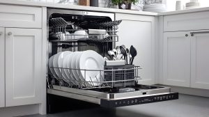 Open dishwasher built into a modern kitchen with white cabinetry, showing clean white plates, bowls, and utensils neatly arranged in the racks. The lower rack holds plates and a utensil basket, while the upper rack contains bowls and smaller items.
