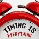 Close-up of a red twin-bell alarm clock with the phrase "TIMING IS EVERYTHING" printed on its white clock face; "TIMING IS" is in bold black text and "EVERYTHING" is in red capital letters.