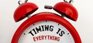 Close-up of a red twin-bell alarm clock with the phrase "TIMING IS EVERYTHING" printed on its white clock face; "TIMING IS" is in bold black text and "EVERYTHING" is in red capital letters.