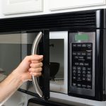 A person opens the door of a mounted stainless steel microwave oven with a curved handle, revealing a dark-colored bowl inside. The microwave has a digital display and a keypad with various cooking options and number buttons. White kitchen cabinets and a decorative glass cabinet are visible in the background.