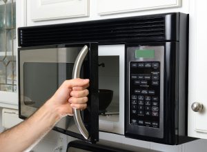 A person opens the door of a mounted stainless steel microwave oven with a curved handle, revealing a dark-colored bowl inside. The microwave has a digital display and a keypad with various cooking options and number buttons. White kitchen cabinets and a decorative glass cabinet are visible in the background.