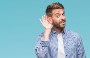 A man with short brown hair and a beard, wearing a blue and white checkered shirt over a white t-shirt, stands against a solid light blue background. He smiles slightly and holds his hand to his ear in a listening gesture, leaning his head slightly forward.