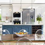 Modern kitchen and dining area with a clean, bright design. The kitchen features white cabinetry, a navy blue island, stainless steel appliances, and pendant lights. A glass dining table with clear acrylic chairs sits in the foreground, topped with a white fruit bowl containing apples and oranges. Potted plants and fresh flowers decorate the space, and large sliding glass doors open to a lush green garden outside. Natural light fills the room through skylights and windows.