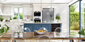 Modern kitchen and dining area with a clean, bright design. The kitchen features white cabinetry, a navy blue island, stainless steel appliances, and pendant lights. A glass dining table with clear acrylic chairs sits in the foreground, topped with a white fruit bowl containing apples and oranges. Potted plants and fresh flowers decorate the space, and large sliding glass doors open to a lush green garden outside. Natural light fills the room through skylights and windows.