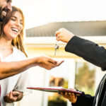 Smiling real estate agent handing house keys to a happy young couple standing outside their new home, with a clipboard of documents in hand and the sun shining in the background.
