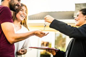 Smiling real estate agent handing house keys to a happy young couple standing outside their new home, with a clipboard of documents in hand and the sun shining in the background.