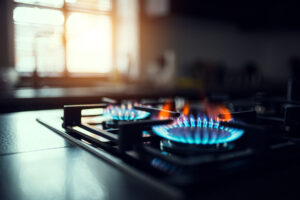 Close-up of a gas stove burner with a blue flame, in a modern kitchen setting. Sunlight streams through a window in the blurred background, creating a warm and inviting atmosphere.