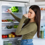A young woman standing in front of an open refrigerator, pinching her nose with a disgusted expression, suggesting a bad smell coming from inside. The fridge shelves are stocked with various foods, including fresh vegetables, eggs, bottled drinks, and leafy greens.