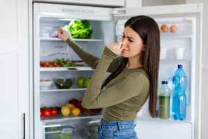 A young woman standing in front of an open refrigerator, pinching her nose with a disgusted expression, suggesting a bad smell coming from inside. The fridge shelves are stocked with various foods, including fresh vegetables, eggs, bottled drinks, and leafy greens.