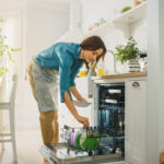 Woman in a bright kitchen loading dishes into a built-in dishwasher, wearing a blue shirt and apron.