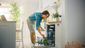Woman in a bright kitchen loading dishes into a built-in dishwasher, wearing a blue shirt and apron.